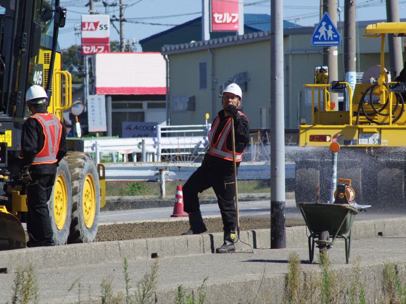 石川県の舗装工事現場で、重機を背景に真剣な表情を見せるロードワークス（白山市）の先輩社員。プロの技術者として活躍（求人）
